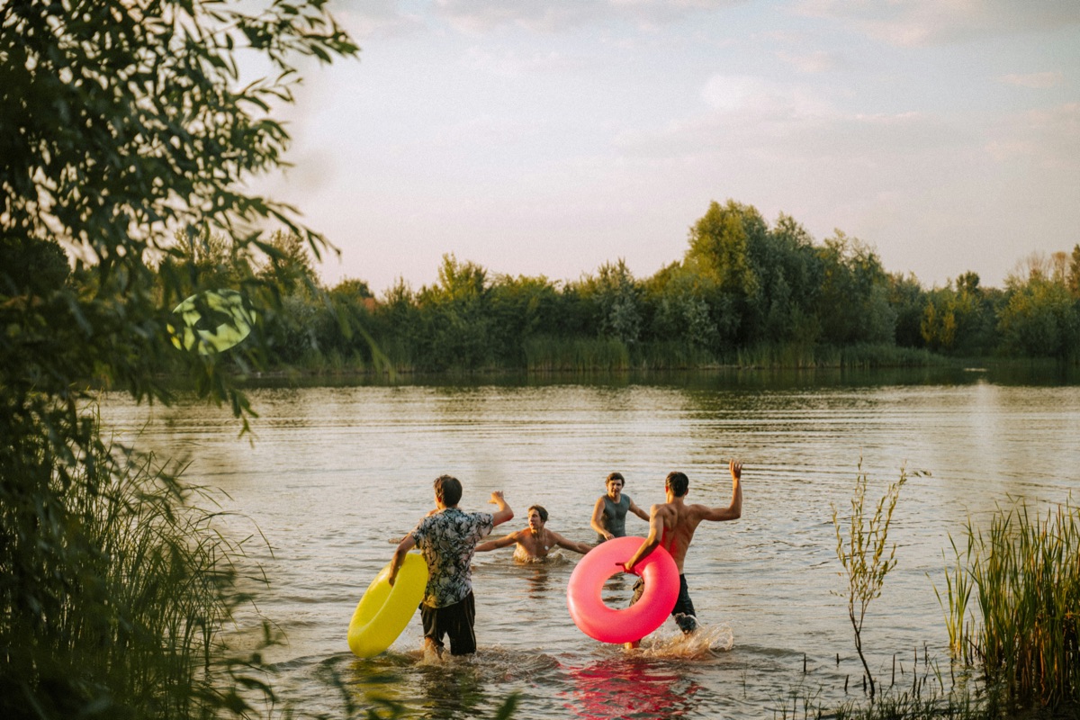 Four friends wading into a lake at golden hour, two carrying inflatable rings